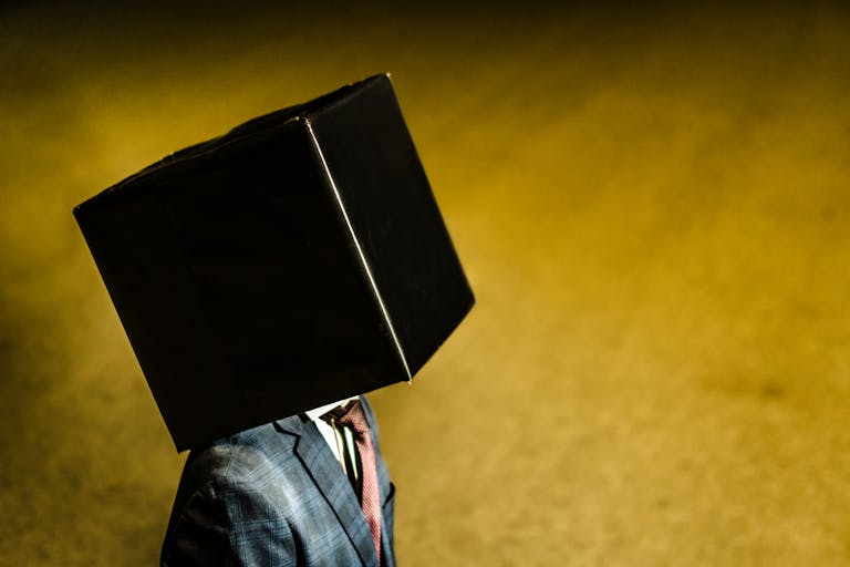 A person in a suit jacket stands outdoors with a black box on their head against a yellow background.