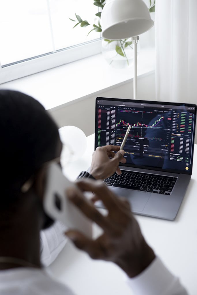 A businessman using a smartphone while analyzing stock market graphs on a laptop indoors.